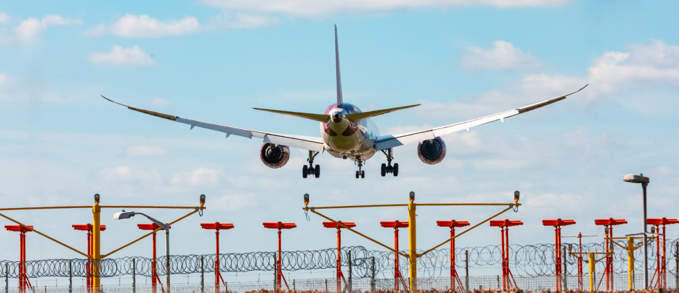Passenger jet descending toward the runway with landing gear down, seen from behind a fence with yellow and red posts and barbed wire.