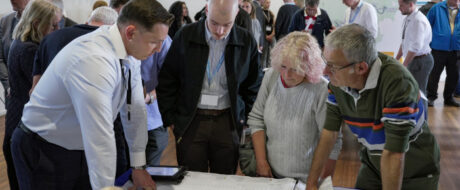 A group of professionals gathered around a table, studying a large document or blueprint laid out for discussion at a conference or workshop.