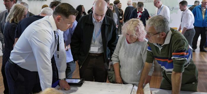 A group of professionals gathered around a table, studying a large document or blueprint laid out for discussion at a conference or workshop.
