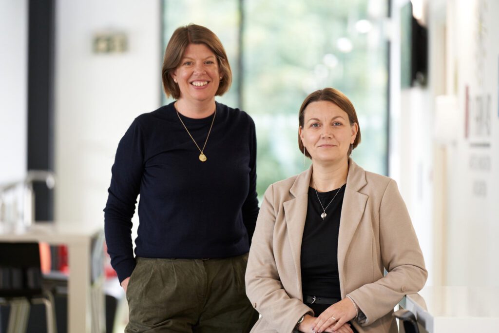 Two women in an office lobby pose for a photo: one standing in a dark blue sweater, the other seated in a beige blazer by a glass railing.