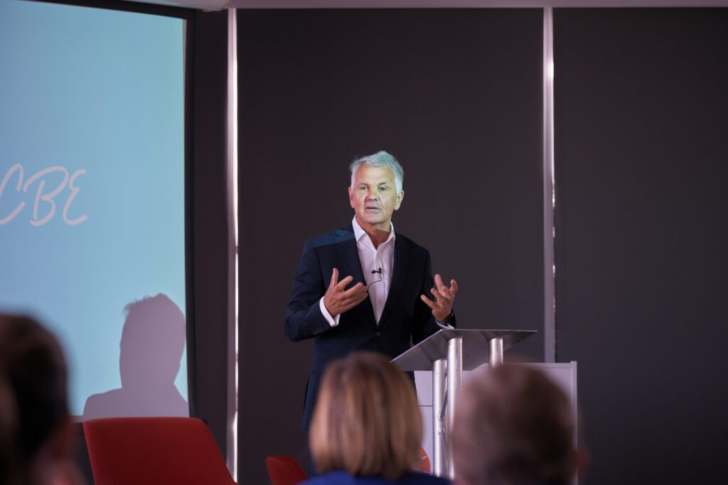 Man in a dark suit delivers a talk from a clear lectern on a stage, audience visible in foreground.