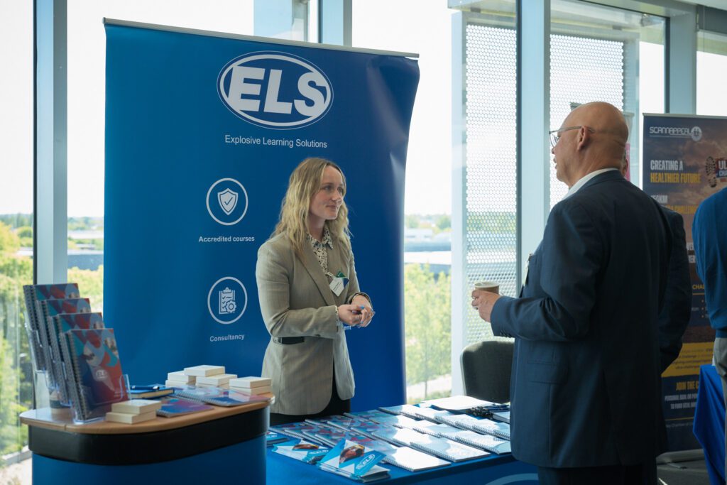 A professional woman standing at a blue ELS booth, speaking to a visitor in a suit near a display of brochures and stacks of blocks.