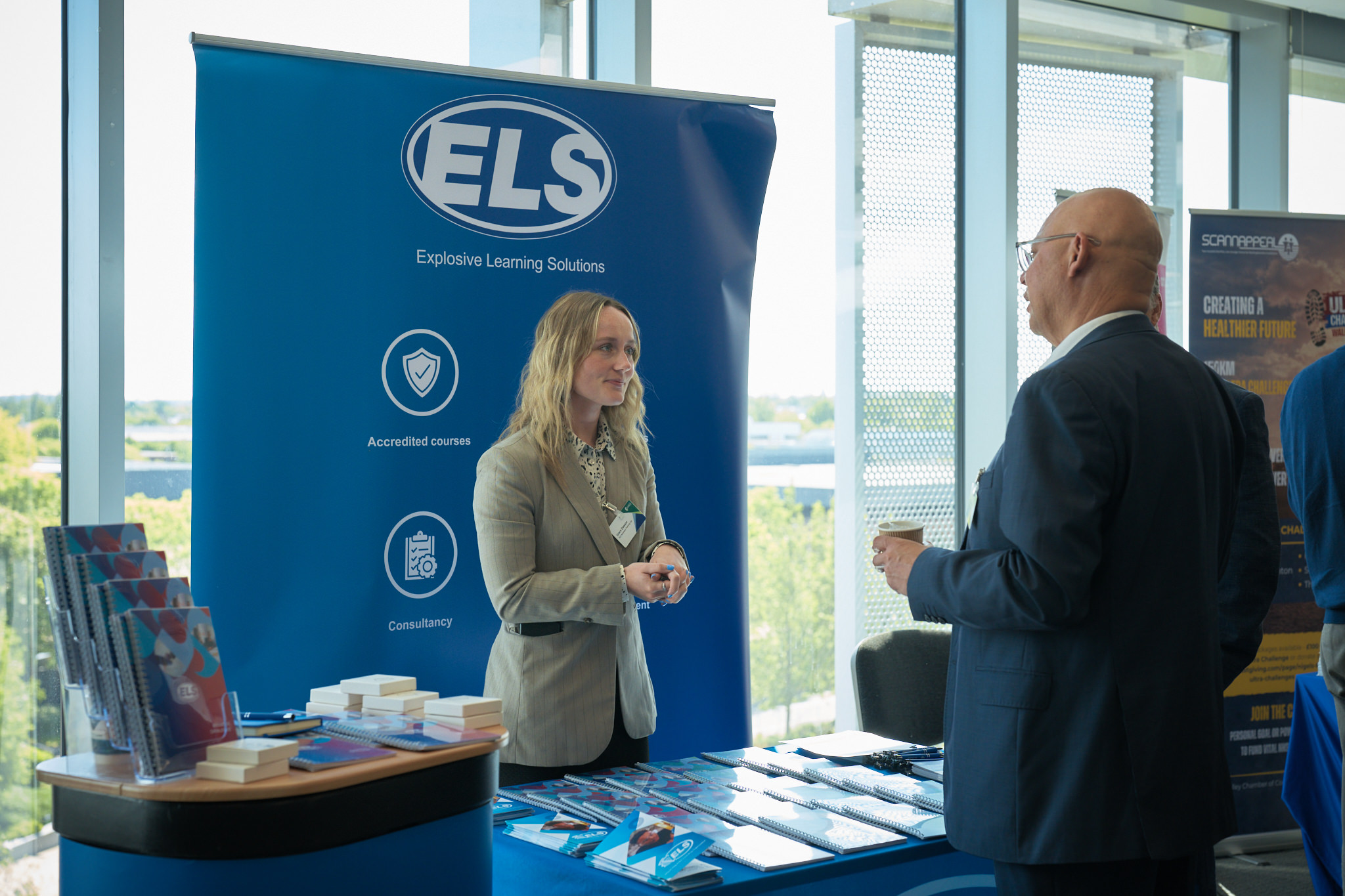 A professional woman standing at a blue ELS booth, speaking to a visitor in a suit near a display of brochures and stacks of blocks.