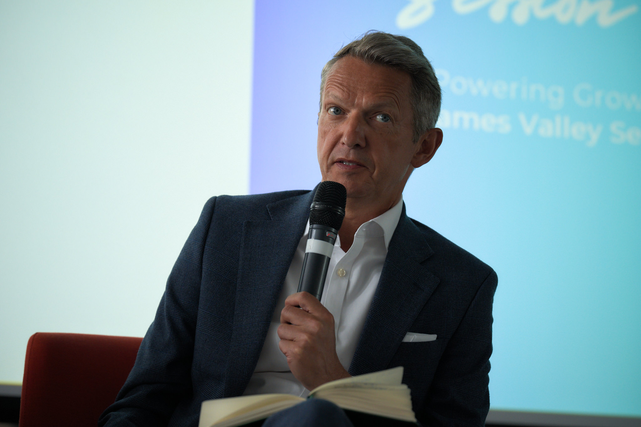 Man in a dark blazer speaks into a handheld microphone, looking to the side, with notes in his lap at a conference stage.