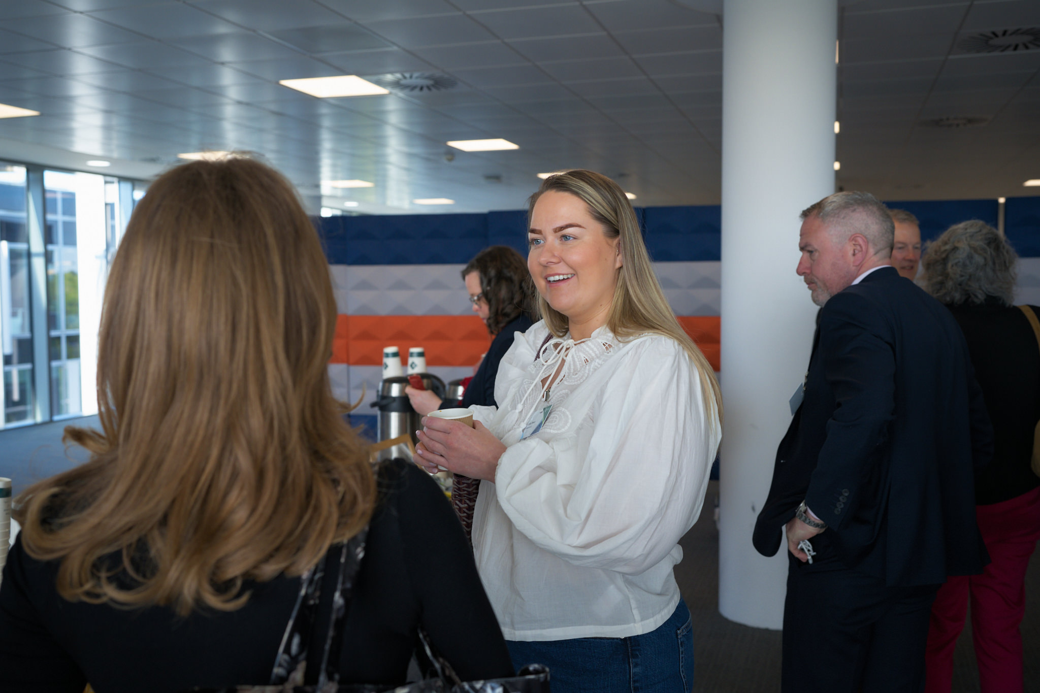 Smiling woman in a white blouse holding a cup chats with colleagues at a bright corporate reception area.