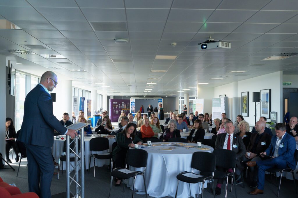 Speaker in a navy suit presenting at a lectern to a large audience in a conference room with round tables and banners in the background.