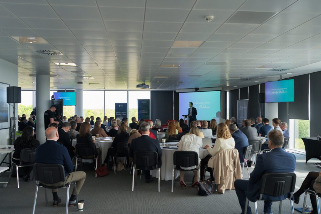 Conference room filled with attendees listening to a speaker at a podium, with large presentation screens in the background.