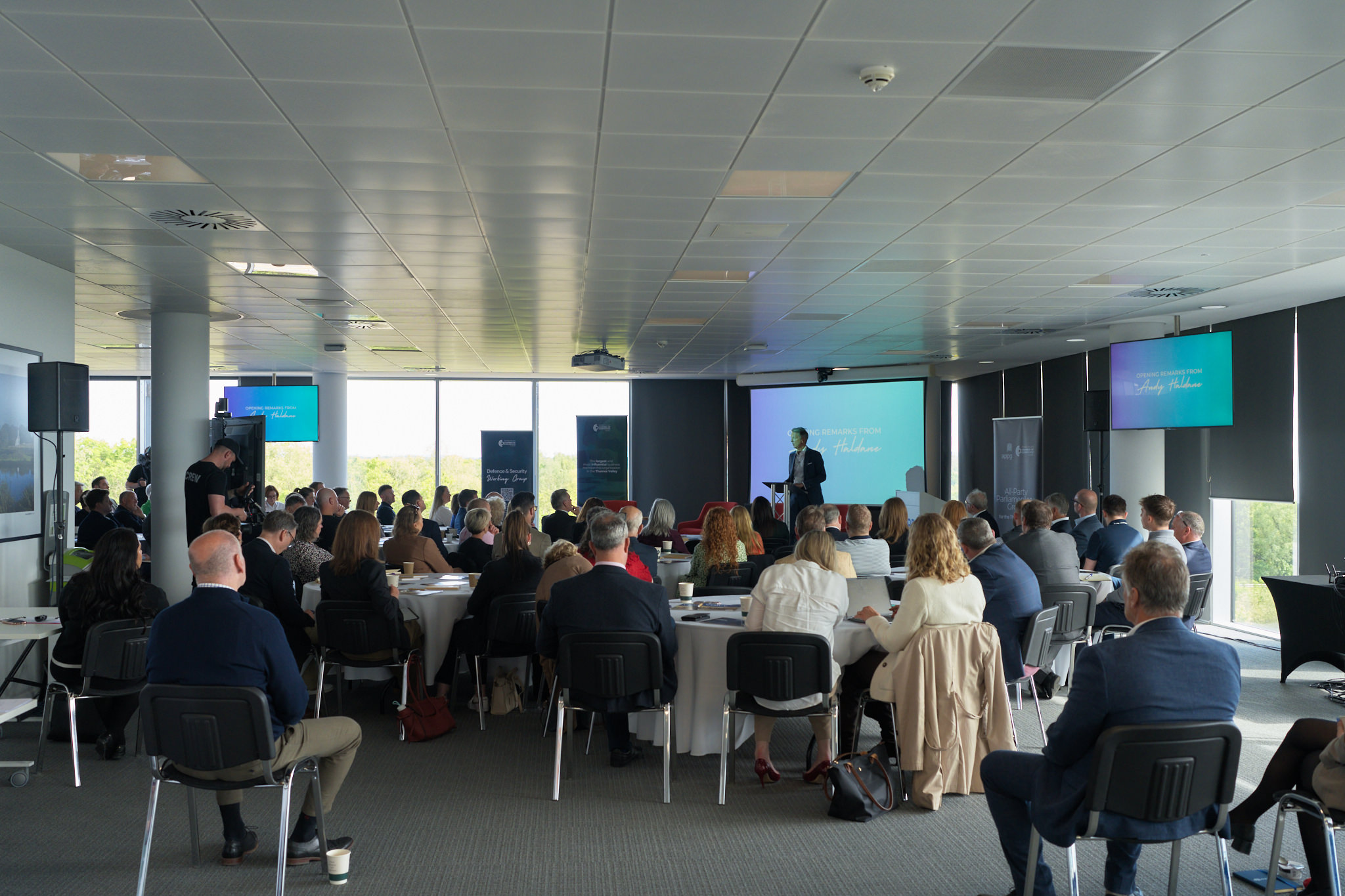 Conference room filled with attendees listening to a speaker at a podium, with large presentation screens in the background.