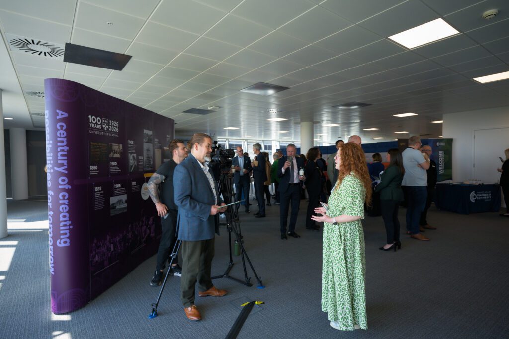 Conference attendees in a lobby near a purple exhibition wall; a camera crew films a man in a blazer while a woman in a green dress talks nearby.