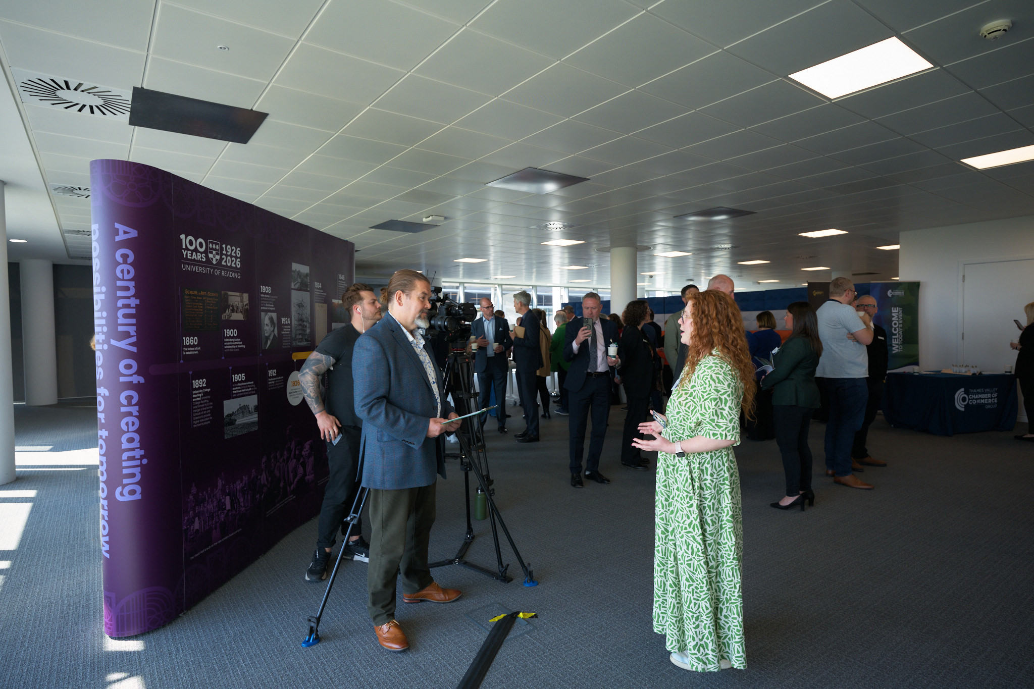 Conference attendees in a lobby near a purple exhibition wall; a camera crew films a man in a blazer while a woman in a green dress talks nearby.