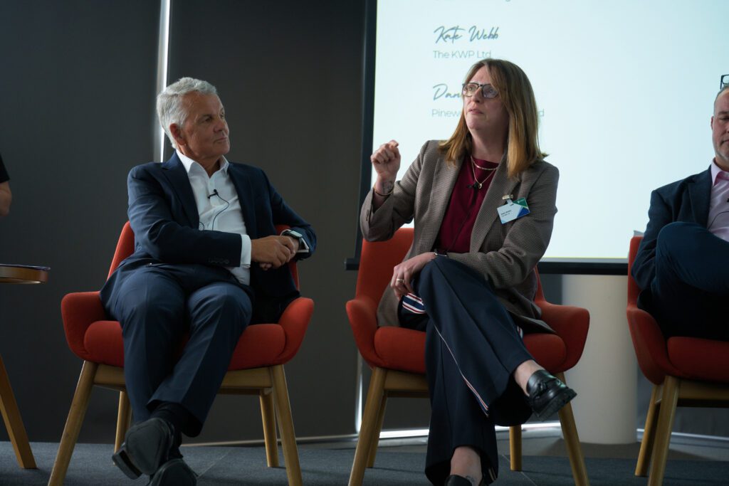 Panel discussion: three professionals seated in red chairs on a stage, one woman speaking and gesturing while the others listen, microphones clipped to their clothing.