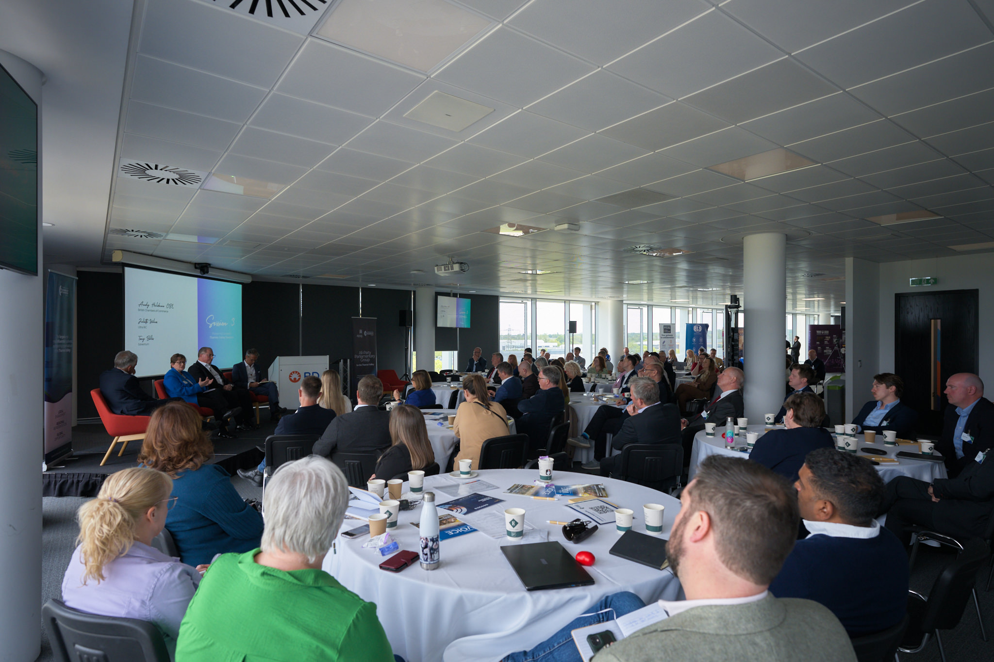 Attendees seated at round tables in a conference room, facing speakers at a panel with a large screen at the front.