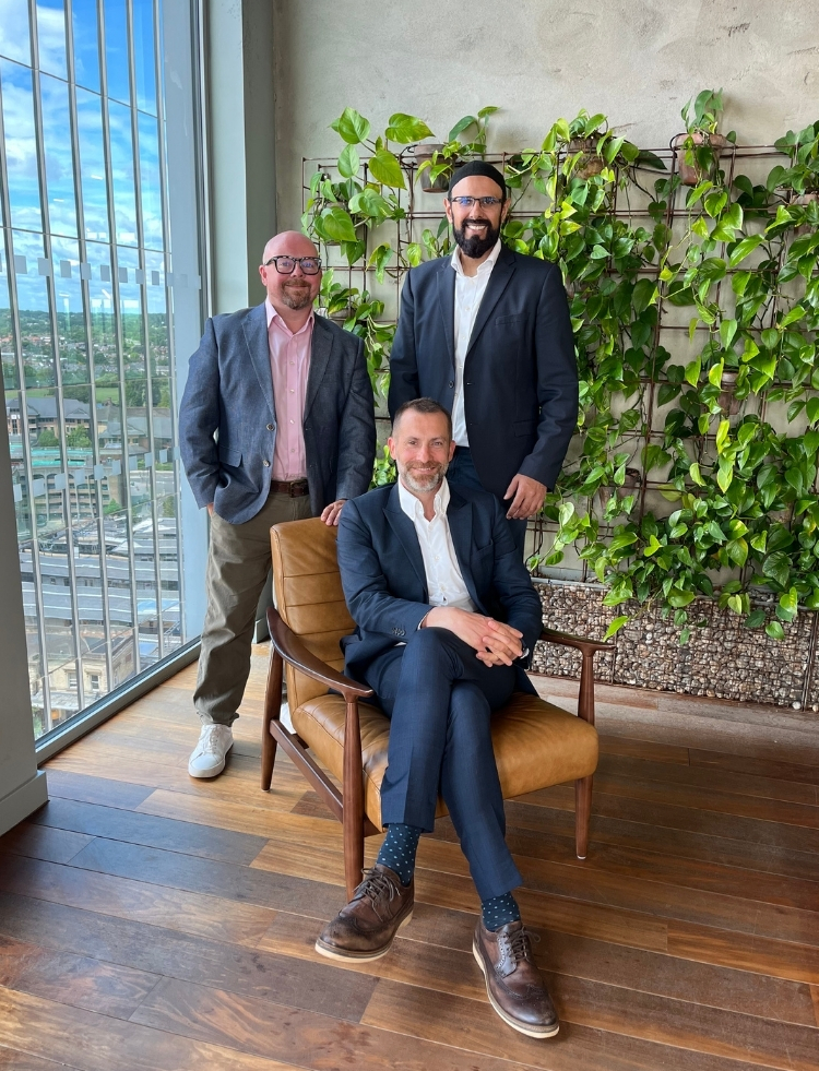Three men in suits pose in a bright office with a green leafy wall; one sits in a brown leather chair while two stand behind him on a wooden floor by a windowed wall.