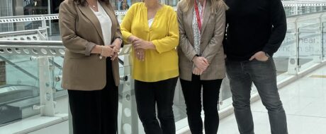 Four adults stand together in a bright indoor shopping center atrium, smiling at the camera with storefronts in the background on a glass railing behind them.