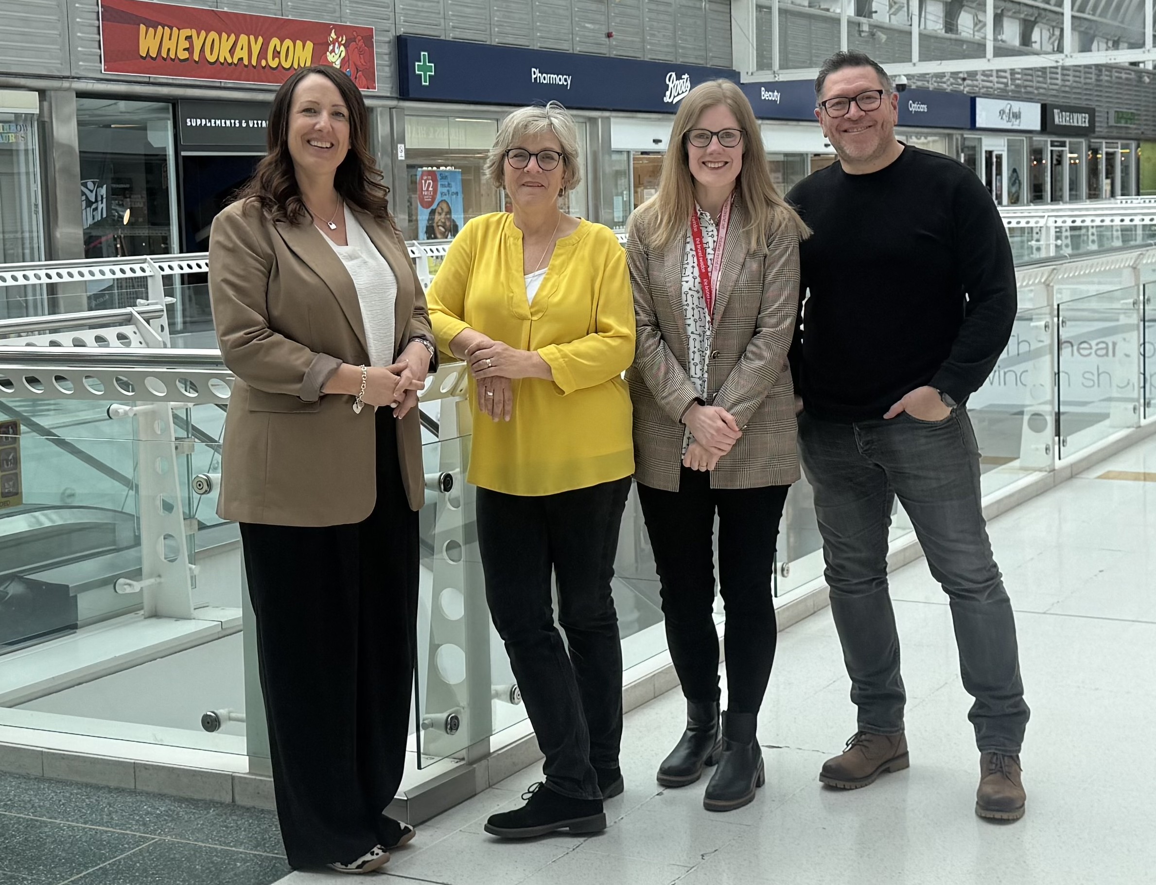 Four adults stand together in a bright indoor shopping center atrium, smiling at the camera with storefronts in the background on a glass railing behind them.