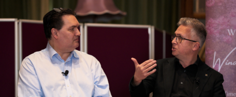 Two men in a panel discussion, each with a lapel mic, speaking to each other on a stage with purple dividers behind them.