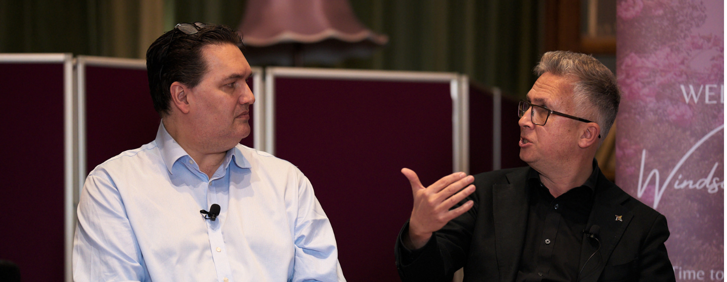 Two men in a panel discussion, each with a lapel mic, speaking to each other on a stage with purple dividers behind them.