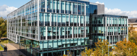 Modern glass office building with vertical and horizontal metal frames under a blue sky, reflecting surroundings in the façade.