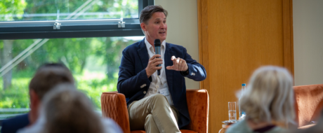 Man in a navy blazer speaks into a handheld microphone while seated on an orange chair during a panel discussion in a bright room with a large window behind him.