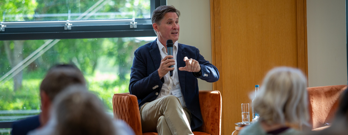 Man in a navy blazer speaks into a handheld microphone while seated on an orange chair during a panel discussion in a bright room with a large window behind him.