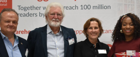 Four adults stand together in front of a banner that reads “Together we reach 100 million readers by …,” smiling for a group photo at a conference or event.