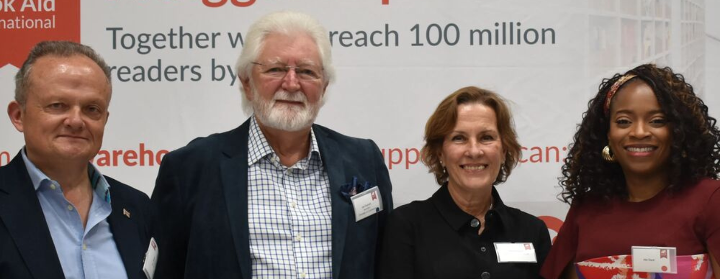 Four adults stand together in front of a banner that reads “Together we reach 100 million readers by …,” smiling for a group photo at a conference or event.