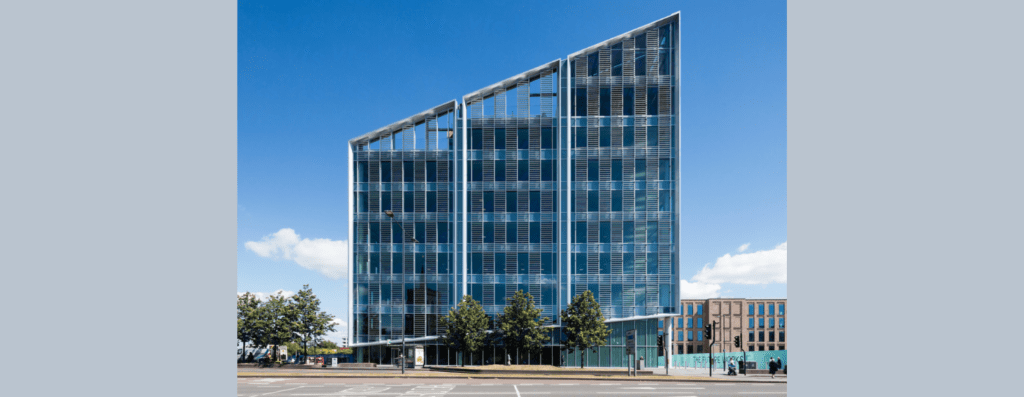 Modern glass office building with three vertical sections along a tree-lined street under a blue sky.