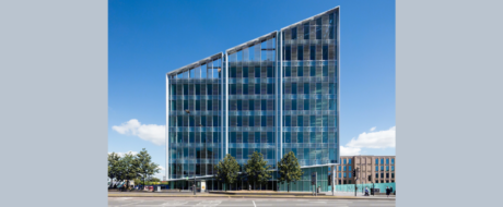 Modern glass office building with three vertical sections along a tree-lined street under a blue sky.