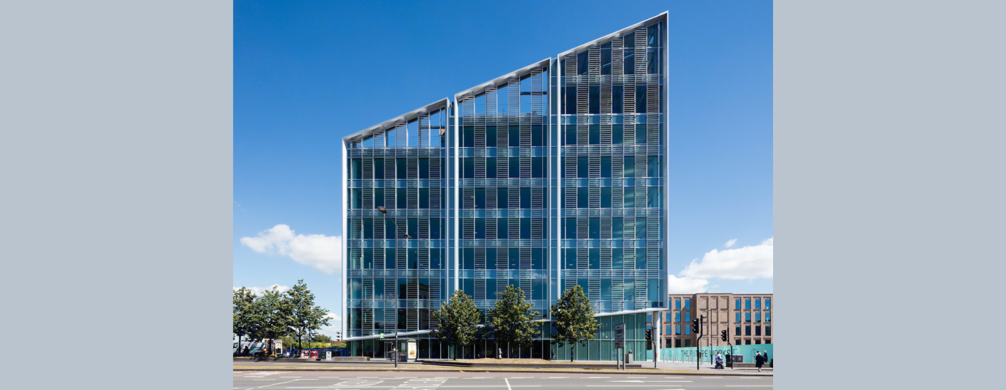 Modern glass office building with three vertical sections along a tree-lined street under a blue sky.