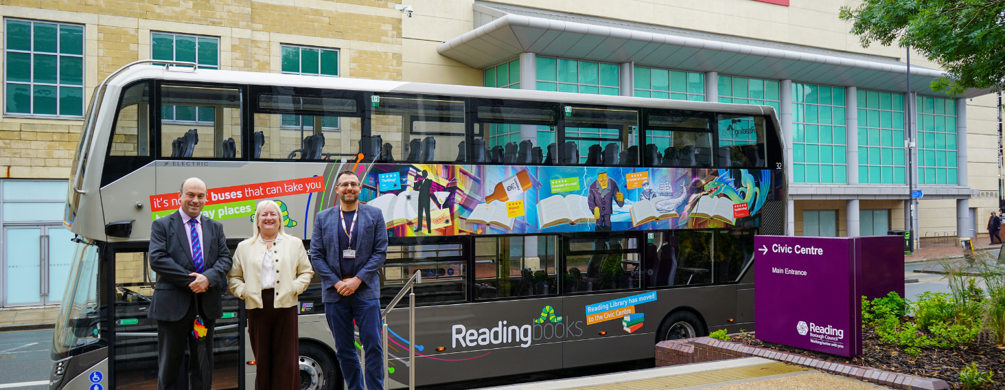 Three people in business attire pose beside a colorful Reading Books library bus outside a civic centre.','A double-decker library bus with vibrant banners reads 'Reading books' parked near a Civic Centre sign; three adults smile.','Library bus with mural advertising books stands in front of a modern building; three people pose for photo.','Purple Civic Centre sign and a modern Reading library bus with murals sit in a plaza, with three adults in front.