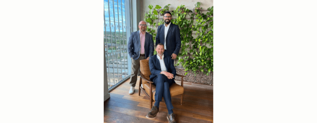 Three men in business attire pose in a plant-filled office; seated man in a navy suit on a brown chair, with two colleagues standing behind him.