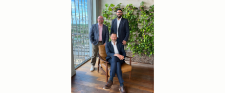 Three men in business attire pose in a plant-filled office; seated man in a navy suit on a brown chair, with two colleagues standing behind him.