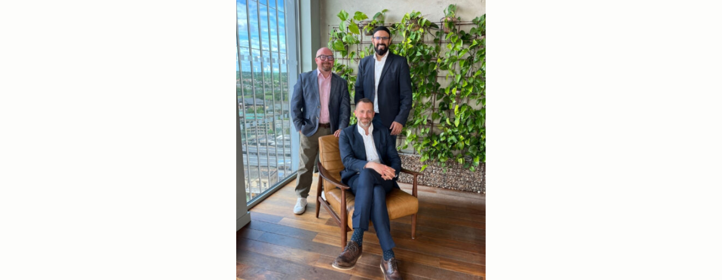 Three men in business attire pose in a plant-filled office; seated man in a navy suit on a brown chair, with two colleagues standing behind him.
