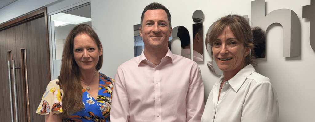 Three colleagues stand together in an office hallway, smiling at the camera—two women on either side of a man in a pink dress shirt, with a company sign on the wall behind them.