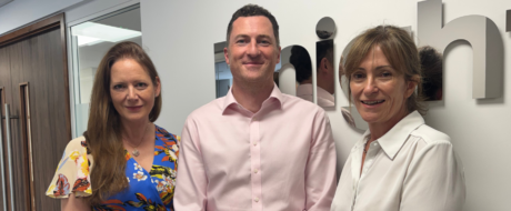 Three colleagues stand together in an office hallway, smiling at the camera—two women on either side of a man in a pink dress shirt, with a company sign on the wall behind them.
