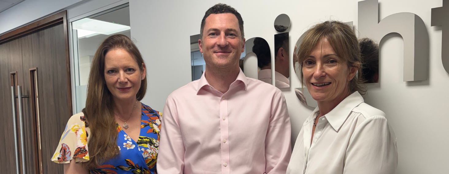 Three colleagues stand together in an office hallway, smiling at the camera—two women on either side of a man in a pink dress shirt, with a company sign on the wall behind them.
