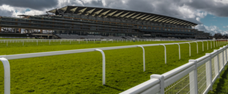 Racing grandstand with tiered seating, white rails, and a green track field under a cloudy sky.
