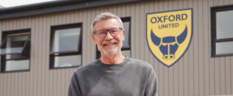Smiling man with gray hair and glasses standing in front of a building with an Oxford United logo behind him.