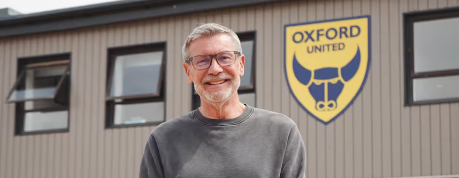 Smiling man with gray hair and glasses standing in front of a building with an Oxford United logo behind him.
