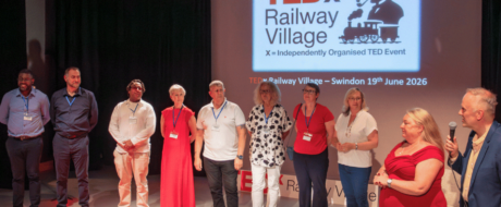 Ten people stand in a line on a stage, facing the audience, with a TEDx Railway Village Swindon projection in the background showing the event logo and date.