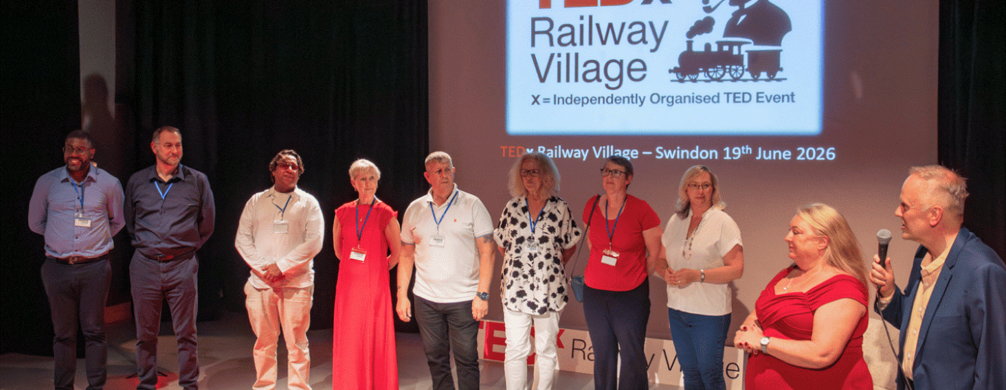 Ten people stand in a line on a stage, facing the audience, with a TEDx Railway Village Swindon projection in the background showing the event logo and date.