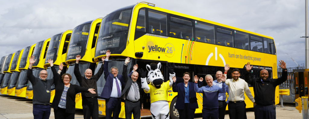 Group of people and a mascot posing with arms raised in front of a line of bright yellow electric buses.",