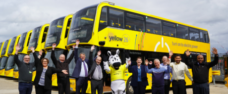 Group of people and a mascot posing with arms raised in front of a line of bright yellow electric buses.",