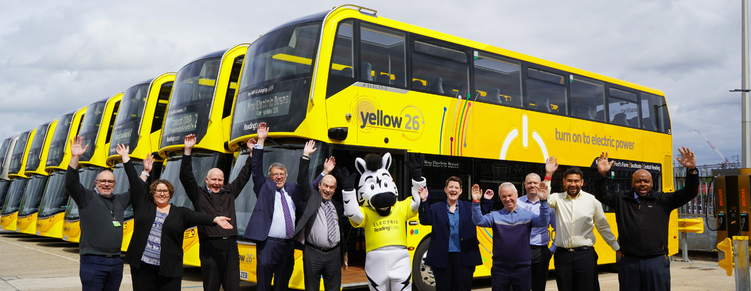 Group of people and a mascot posing with arms raised in front of a line of bright yellow electric buses.",