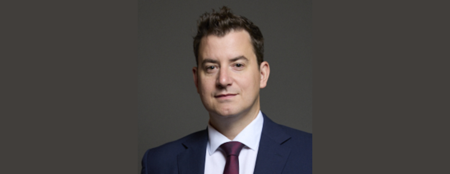 Head-and-shoulders portrait of a man in a navy suit, white shirt, burgundy tie, looking at the camera against a gray background