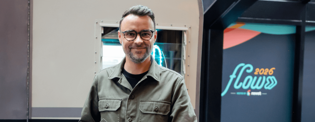 Smiling man with glasses and short gray beard wearing an olive-green jacket, standing in an industrial setting near a metal machine, with a colorful display that reads 'flow 2026' in the background