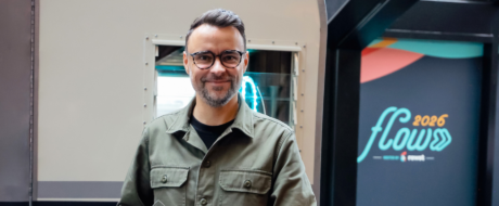 Smiling man with glasses and short gray beard wearing an olive-green jacket, standing in an industrial setting near a metal machine, with a colorful display that reads 'flow 2026' in the background
