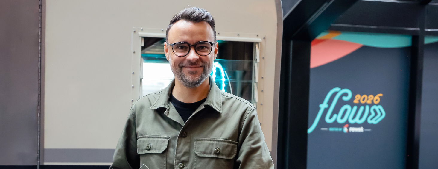 Smiling man with glasses and short gray beard wearing an olive-green jacket, standing in an industrial setting near a metal machine, with a colorful display that reads 'flow 2026' in the background