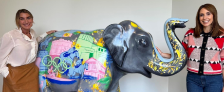 Two smiling women pose beside a large gray elephant sculpture painted with colorful city scenes and flowers inside a bright room, likely a gallery.