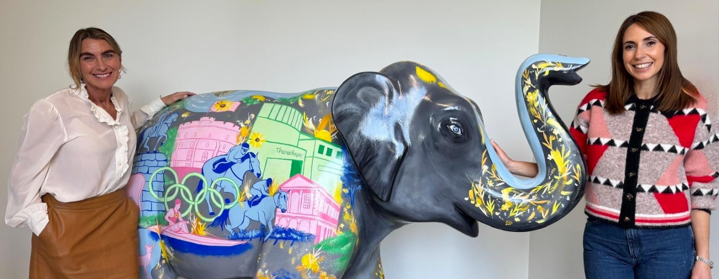 Two smiling women pose beside a large gray elephant sculpture painted with colorful city scenes and flowers inside a bright room, likely a gallery.